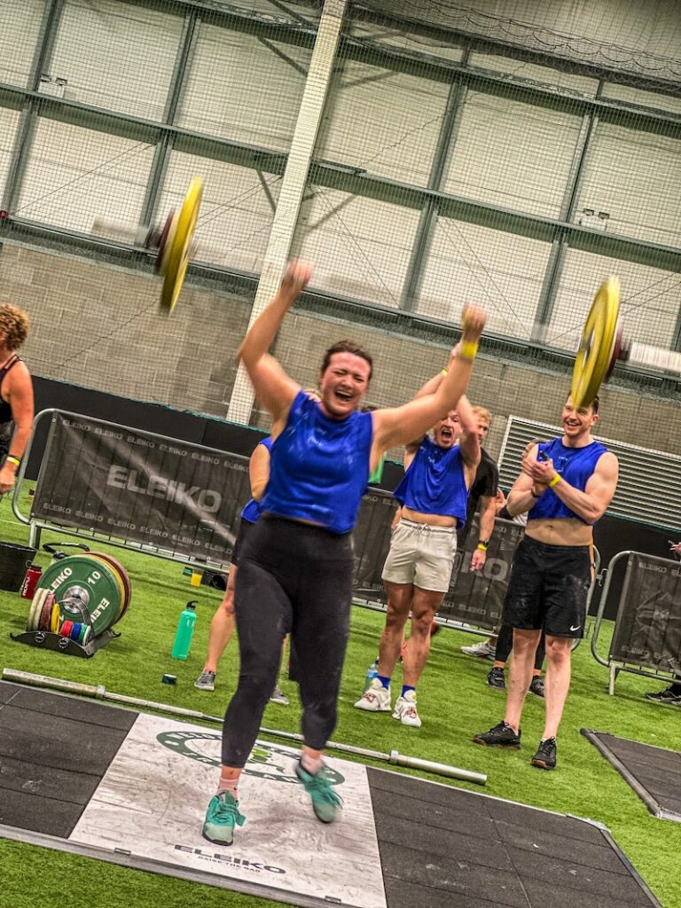 A dynamic gym scene capturing the three pillars: a skilled coach in the foreground guiding a diverse group through a functional workout, with athletes of all levels collaborating on Olympic lifts and gymnastics near premium Eleiko bars and Rogue rigs, all under bright lighting that highlights the energetic, welcoming community vibe.

